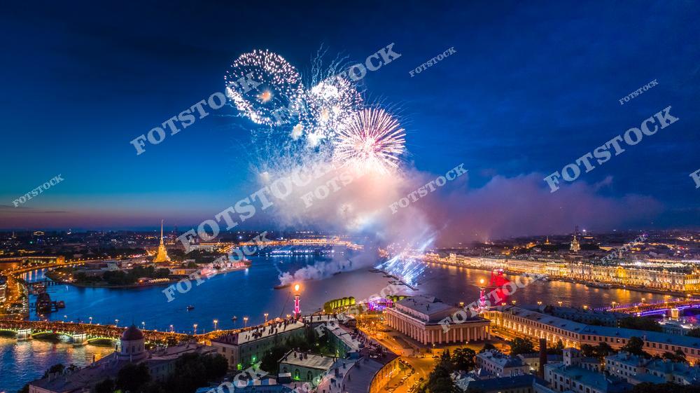 На фотографии изображен фейерверк над центральной частью Санкт-Петербурга, с видом на Неву и Стрелку Васильевского острова. Яркие вспышки салюта озаряют ночное небо и отражаются в водах реки, создавая впечатляющее зрелище. На переднем плане можно увидеть здание Биржи, Ростральные колонны и Петропавловскую крепость, что делает этот кадр символичным для северной столицы. Такое событие привлекает тысячи зрителей и символизирует торжество и красоту Петербурга.